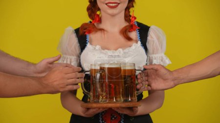 Foto de Primer plano fondo amarillo aislado video de una joven alemana, camarera con un traje tradicional, sosteniendo un plato con cerveza, cerveza. Las manos quitan la cerveza del plato. Octoberfest, anuncio. - Imagen libre de derechos