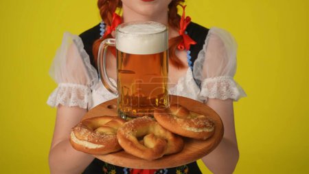 Téléchargez les photos : Gros plan fond jaune plan isolé d'une jeune Allemande méconnaissable, serveuse vêtue d'un costume traditionnel, tenant un plateau de bretzels et un verre de bière. Octoberfest - en image libre de droit