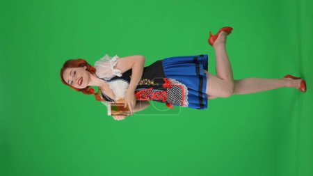 Foto de Pantalla verde vertical de tamaño completo aislado croma clave disparo de una joven mujer alemana, camarera, sosteniendo un vaso de cerveza, cerveza, coqueteando, levantando la pierna. Festival de octubre, festival de la cerveza, publicidad. - Imagen libre de derechos