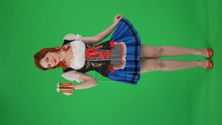 Foto de Pantalla verde vertical de tamaño completo aislado croma clave disparo de una joven alemana, camarera, sosteniendo un vaso de cerveza, cerveza, coqueteando, tirando de su falda. Festival de octubre, festival de la cerveza, publicidad. - Imagen libre de derechos