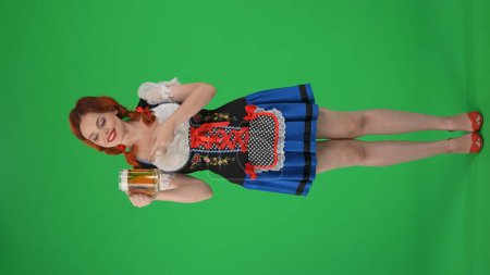 Foto de Pantalla verde vertical de tamaño completo aislado croma clave de una joven mujer alemana, camarera, sosteniendo un vaso de cerveza, cerveza, señalándolo y sonriendo. Festival de octubre, festival de la cerveza, publicidad. - Imagen libre de derechos