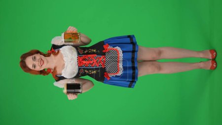 Photo for Full-size vertical green screen isolated chroma key shot of a young German woman, waitress, holding two glasses of lager, light and dark beer, smiling. Octoberfest, beer festival, advertisement. - Royalty Free Image