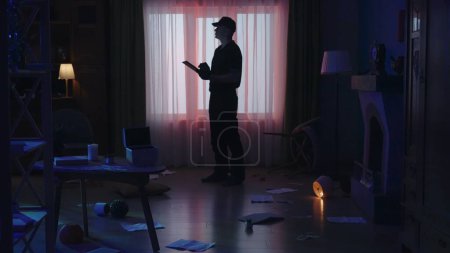 Photo for Crime scene creative concept. Police man with tablet standing in the dark living room. Portrait of male in uniform in the apartment with signs of robbery. - Royalty Free Image
