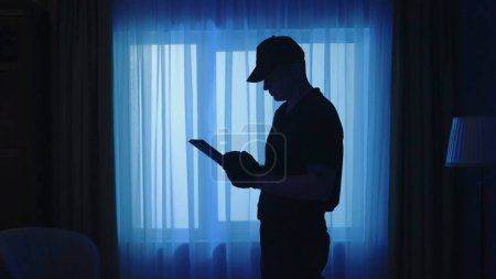 Photo for Crime scene creative concept. Policeman with tablet in the dark living room. Portrait of a man in uniform in the apartment writing something, looking around. - Royalty Free Image