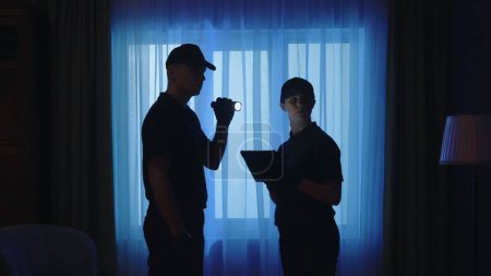 Téléchargez les photos : Concept créatif de scène de crime. Des policiers debout dans la chambre noire. Portrait d'un homme et d'une femme avec tablette et lampe de poche, regardant autour de l'endroit. - en image libre de droit