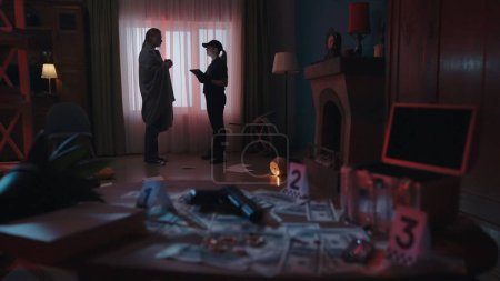 Crime scene creative concept. Young woman in blanket talking with police officer in the dark room, signs of robbery. Table with evidence at the front.
