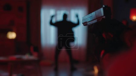 Foto de El arresto de un ladrón en la escena del crimen, en un apartamento oscuro iluminado por la luz roja. El delincuente está de pie con los brazos levantados, la espalda a la cámara. Un arma en la mano de un policía en una borrosa - Imagen libre de derechos