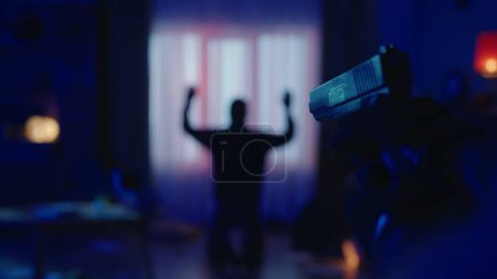 Photo for The arrest of a burglar at the crime scene, in a dark apartment lit by blue light. The offender is on his knees, with his back to the camera with his hands up. A gun in the hand of a policeman on a - Royalty Free Image