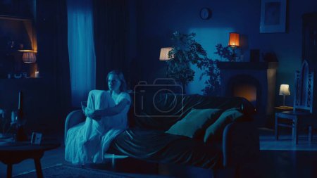 Photo for Shot of a young woman sitting on the couch, embracing, hugging her legs and mumbling, whispering something as if she is posessed by a ghost, demon, poltergeist. Paranormal activity, supernatural. - Royalty Free Image