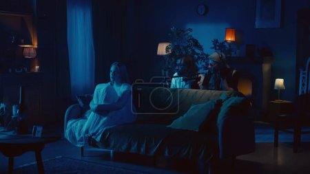 Photo for Shot of two ghost hunters raising from behind the couch trying to capture a posessed female, ghost, poltergeist sitting on the couch, mumbling, whispering something on video. Paranormal activity. - Royalty Free Image