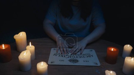 Photo for Shot capturing a womans figure having an ouija board session, seance, surrounded by candles in the dark. She holds a planchette the board. Ominious, paranormal activity, supernatural. - Royalty Free Image