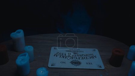 Photo for Shot capturing a a table with an ouija board and a planchette on top. Session, seance. Board is surrounded by candles in the dark. Ominious atmosphere, paranormal activity, supernatural. - Royalty Free Image