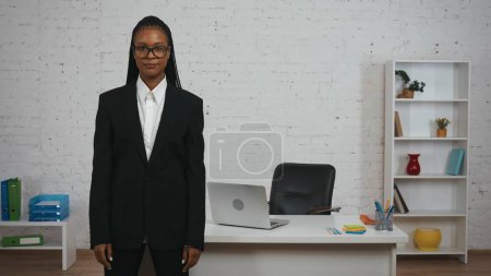 Photo for Modern businesswoman creative concept. Portrait of young African American female in the office. Woman in black suit and glasses standing in front of the desk looking at the camera. - Royalty Free Image