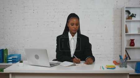 Foto de Concepto creativo de mujer de negocios moderna. Retrato de una joven afroamericana en la oficina. Mujer con traje negro sentada en el escritorio, escribiendo en cuaderno. - Imagen libre de derechos