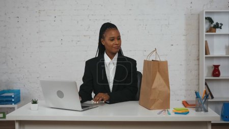 Téléchargez les photos : Concept créatif femme d'affaires moderne. Portrait de jeune femme afro-américaine au bureau. Femme assise au bureau travaillant, regardant et souriant au sac en papier sur la table. - en image libre de droit