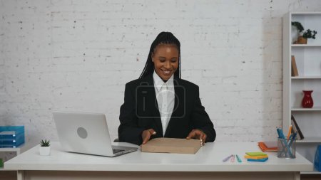 Foto de Concepto creativo de mujer de negocios moderna. Retrato de una joven afroamericana en la oficina. Mujer en el escritorio sonriendo expresión de cara feliz sosteniendo la caja de pizza, orden de la hora del almuerzo. - Imagen libre de derechos