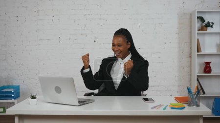 Photo for Modern businesswoman concept. Portrait of young African American female in the office. Woman sitting at the desk smiling happily throwing hands up in the air, pumping fists. - Royalty Free Image