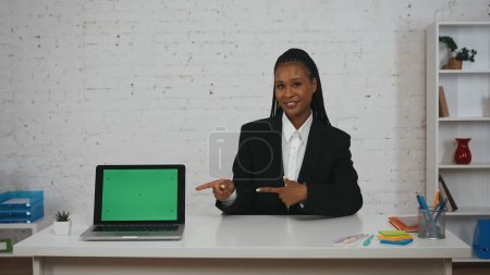 Téléchargez les photos : Concept de femme d'affaires moderne. Portrait de jeune femme afro-américaine au bureau. Femme assise au bureau souriant, pointant vers l'ordinateur portable. Espace publicitaire, maquette de l'espace de travail. - en image libre de droit