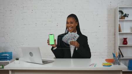 Téléchargez les photos : Concept de femme d'affaires moderne. Portrait de jeune femme afro-américaine au bureau. Femme tenant smartphone et argent, souriant en regardant la caméra. Espace publicitaire, maquette de l'espace de travail. - en image libre de droit
