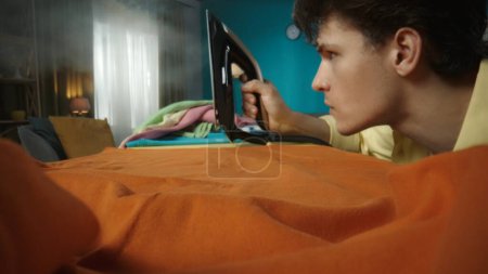 Photo for Close up indoor shot of a young brunette mans face. The man is using a steam iron to iron his clothes. She looks focused. Household appliances, chores. - Royalty Free Image