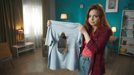 Photo for Middle shot of a young redhead woman holding a t-shirt that was burnt with an iron. The woman looks confused and sorry about the accident. Household appliances, chores, advertisement. - Royalty Free Image
