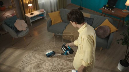 Photo for Shot from above of a young brunette man vacuuming the carpet in his living room, testing, using new vacuum cleaner, product. Indoor, apartment scene. Household appliances, chores. Advertisement. - Royalty Free Image