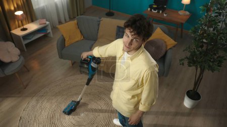 Photo for Shot from above, joyful, happy young man vacuuming the carpet in his living room, looking at the camera and smiling. Household appliances, chores. Advertisement. Indoor, apartment scene. Household - Royalty Free Image