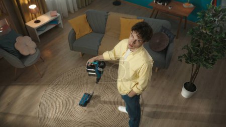 Photo for Shot from above, tired, exhausted young man vacuuming the carpet in his living room, looking at the camera with a disappointed, dissatisfied look. Household appliances, chores. Advertisement. - Royalty Free Image