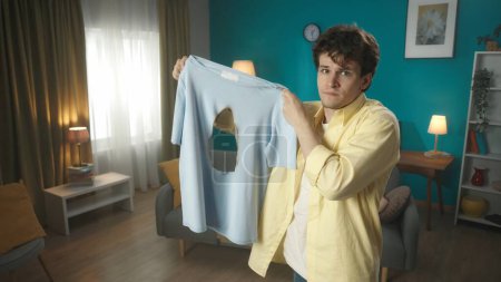 Photo for Middle shot of a young brunette man holding a t-shirt that was burnt with an iron. The man looks confused and sorry about the accident. Household appliances, chores, advertisement. - Royalty Free Image