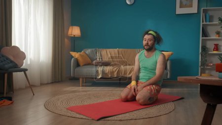 Photo for A middle-aged man sits on a fitness mat and looks at the camera with a sad expression on his face. His long hair is pulled back by a brightly colored bandage. Fitness at home. - Royalty Free Image