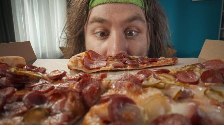 Photo for Middle-aged man with long hair and fitness bandage is shown in a close-up shot of a pizza box against a blue wall. Shot with macro shot Looking at a slice of pizza shows curiosity, interest, surprise. - Royalty Free Image