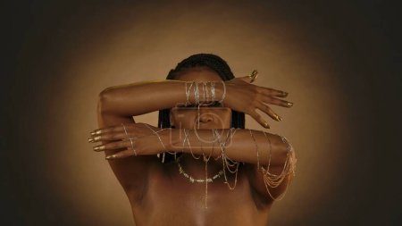 Foto de Una mujer afroamericana demuestra sus manos con brazaletes de cadena dorada, sus yemas de los dedos y las uñas están pintadas con pintura dorada. Mujer Seminude en el estudio sobre un fondo marrón con luz circular - Imagen libre de derechos