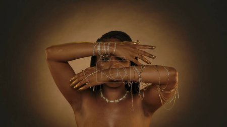 Foto de Una mujer afroamericana demuestra sus manos con brazaletes de cadena dorada, sus yemas de los dedos y las uñas están pintadas con pintura dorada. Mujer Seminude en el estudio sobre un fondo marrón con luz circular - Imagen libre de derechos