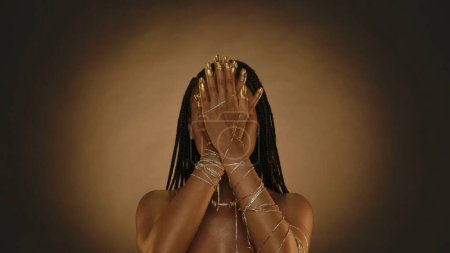 Téléchargez les photos : Femme afro-américaine a couvert son visage avec ses mains dans le studio sur un fond brun avec de la lumière circulaire. Les mains des femmes sont en chaînes d'or, bracelets, bout des doigts et les ongles sont peints avec - en image libre de droit