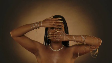 Téléchargez les photos : Femme afro-américaine a couvert son visage avec ses mains, laissant une fente pour ses yeux dans le studio sur un fond brun avec de la lumière circulaire. Les mains des femmes sont en chaînes d'or, bracelets, bout des doigts - en image libre de droit