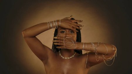 Téléchargez les photos : Femme afro-américaine a couvert son visage avec ses mains, laissant une fente pour ses yeux dans le studio sur un fond brun avec de la lumière circulaire. Les mains des femmes sont en chaînes d'or, bracelets, bout des doigts - en image libre de droit