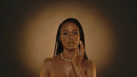 Photo for African American woman applies golden paint on her lips, liquid gold. Seminude woman with gold chain bracelets on her hands, with a necklace around her neck on a brown background with circular light - Royalty Free Image