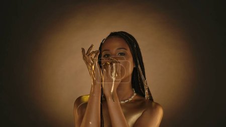 Photo for A woman with golden lips, golden skin and golden chains in her hair in the studio on a brown background with circular light. Arms of a seminude African American woman in liquid gold, or gold paint - Royalty Free Image