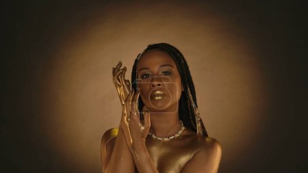 Photo for A woman with golden lips, golden skin and golden chains in her hair in the studio on a brown background with circular light. Arms of a seminude African American woman in liquid gold, or gold paint - Royalty Free Image