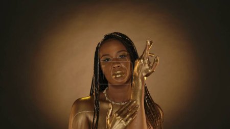 Photo for A woman with golden lips, golden skin, golden chains in her hair on a brown background with circular light. A seminude African American woman applies, rubs golden paint, liquid gold into the skin of - Royalty Free Image