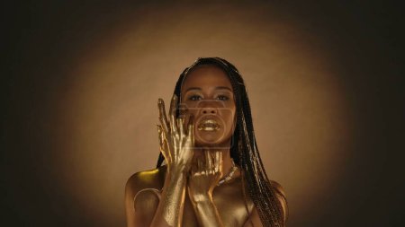 Téléchargez les photos : Une femme aux lèvres dorées, à la peau dorée et aux chaînes dorées dans ses cheveux en studio sur un fond brun avec une lumière circulaire. Armoiries d'une femme afro-américaine séminude en or liquide, ou peinture dorée - en image libre de droit
