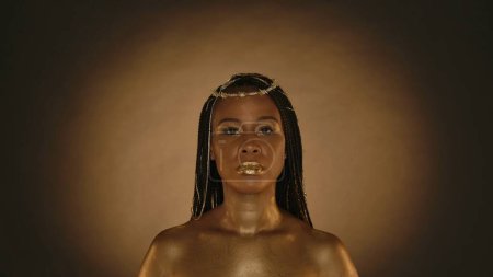 Photo for Portrait of a seminude African American woman in the style of Cleopatra in the studio on a brown background with circular light. A woman with creative golden metallic makeup, golden skin and jewelry - Royalty Free Image