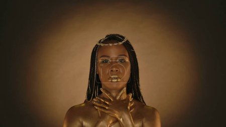 Téléchargez les photos : Portrait d'une femme séminude à la peau dorée et au maquillage métallique doré sur fond brun avec lumière circulaire. La femme enroule ses bras autour de son cou, touche la peau de ses épaules - en image libre de droit