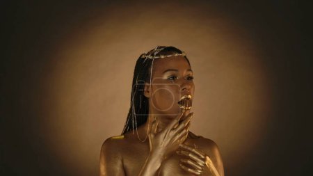 Téléchargez les photos : Femme afro-américaine dans le style Cléopâtre sur un fond brun avec une lumière circulaire. Une femme avec une peau dorée et des bijoux sur la tête tient un bonbon au chocolat doré avec ses lèvres, dans sa bouche - en image libre de droit