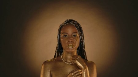 Photo for Portrait of a African American woman with golden drops of tears on her face. Seminude woman with golden metallic make up, golden skin and with jewelry in her hair on a brown background with circular - Royalty Free Image
