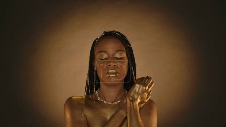 Photo for Portrait of a African American woman in the style of Cleopatra with closed eyes on a brown background with circular light. Woman with creative golden makeup, skin, necklace around her neck, jewelry in - Royalty Free Image