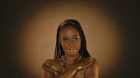 Photo for A seminude African American woman in the style of Cleopatra on a brown background with circular light. Woman with creative golden metallic makeup, golden skin, necklace around her neck and jewelry in - Royalty Free Image