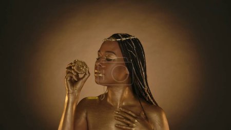 Foto de Mujer afroamericana en estilo Cleopatra sobre fondo marrón con luz circular. Una mujer con la piel en pintura dorada, con joyas en la cabeza, sostiene la mitad de un limón. Diseño de arte de moda - Imagen libre de derechos