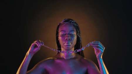 Téléchargez les photos : Une femme afro-américaine met une chaîne autour de son cou, avec des gouttes d'or liquide, dans la lumière rose néon. Femme avec un maquillage doré et des gouttes d'or de larmes sur le visage dans un studio sur un fond brun - en image libre de droit