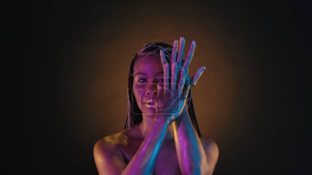 Téléchargez les photos : Une Afro-Américaine montre ses mains en peinture dorée sur fond brun avec une lumière circulaire. Femme avec un maquillage de style Cleopatra créatif et des bijoux en or dans ses cheveux en lumière rose néon - en image libre de droit